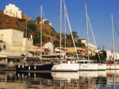 Ιστιοφόρα και σκάφη αγκυροβολημένα στο λιμάνι του Κόρφου στην Κέα, με παραδοσιακά κτίρια στο φόντο.