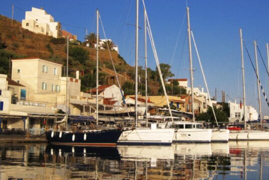 Ιστιοφόρα και σκάφη αγκυροβολημένα στο λιμάνι του Κόρφου στην Κέα, με παραδοσιακά κτίρια στο φόντο.