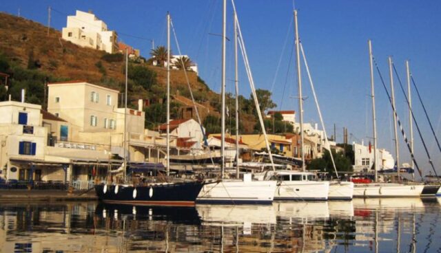 Ιστιοφόρα και σκάφη αγκυροβολημένα στο λιμάνι του Κόρφου στην Κέα, με παραδοσιακά κτίρια στο φόντο.