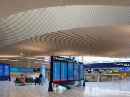 Επιβάτης περιμένει δίπλα σε ψηφιακές οθόνες αναχωρήσεων σε σύγχρονη αίθουσα αεροδρομίου.
